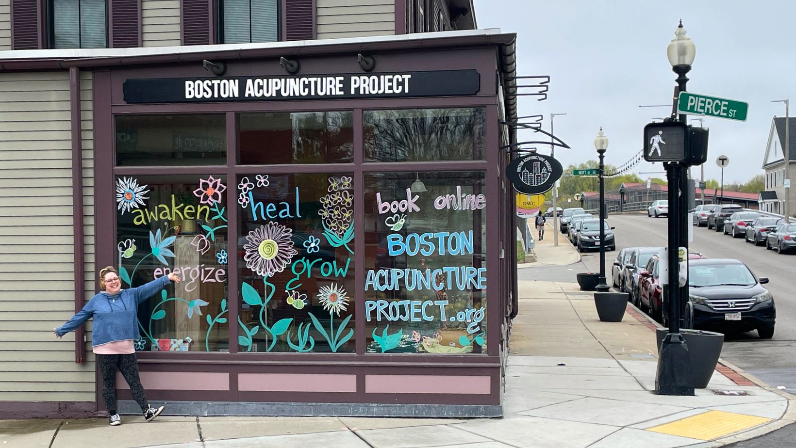 Dee poses in front of the windows at Boston Acupuncture Project which she has painted with flowers and the words awaken, heal, energize, thrive, grow. Book online at Boston Acupuncture Project dot com.
