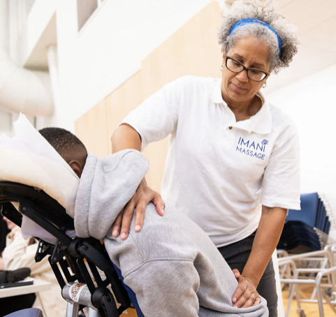 Christine Rose a Black woman in a white t-shirt with white-gray hair in a blue headband gives a chair massage