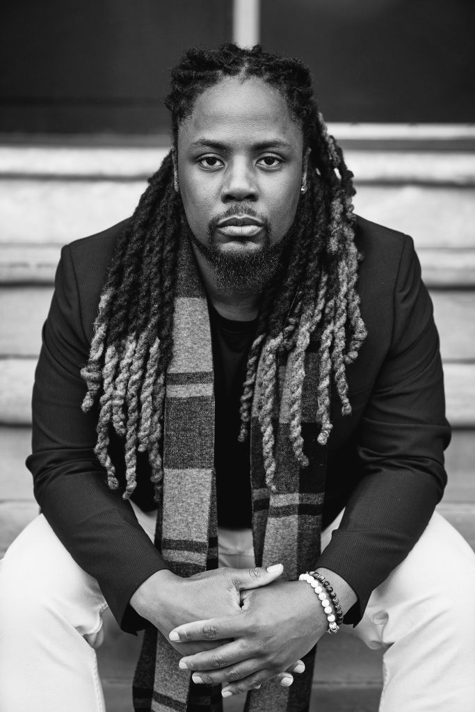 Jonathan, a Black man with locs, gazes into the camera in an artistic black-and-white photographic portrait. 
