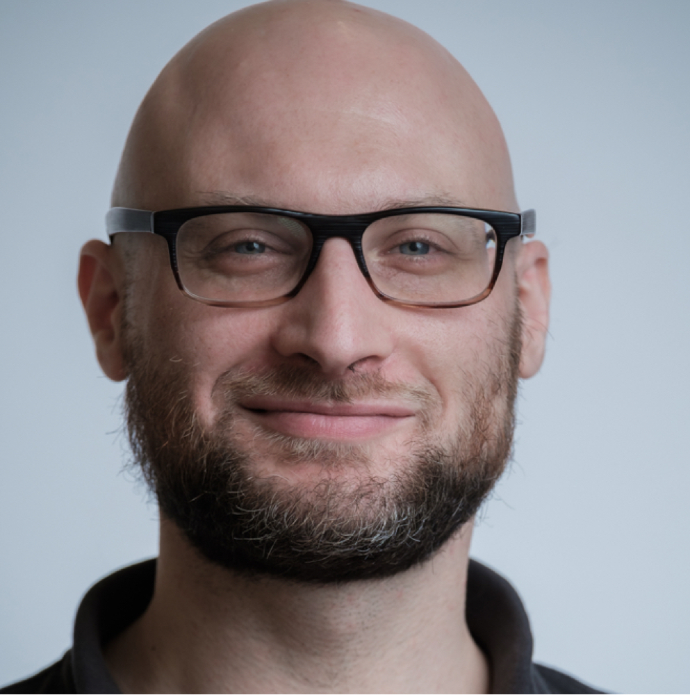 White person with a beard, glasses, and no hair on head smiles at camera